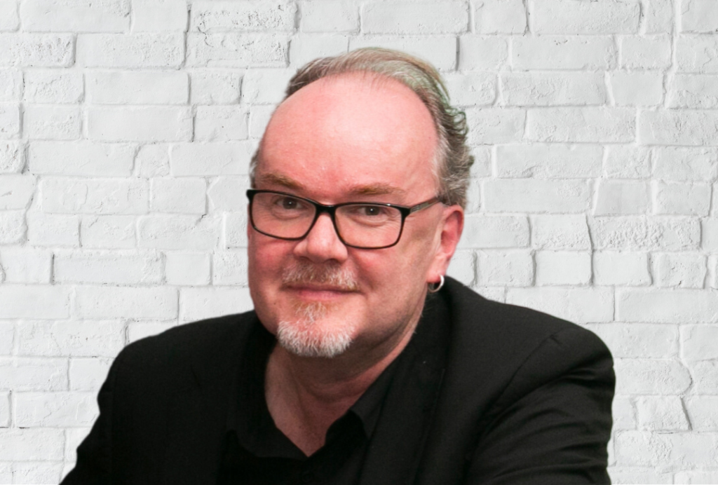 Man smiles at the camera with grey hair and a beard. He wears glasses.