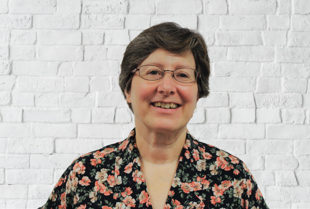 A woman with short hair and glasses smiles at the camera. She is wearing a floral top.