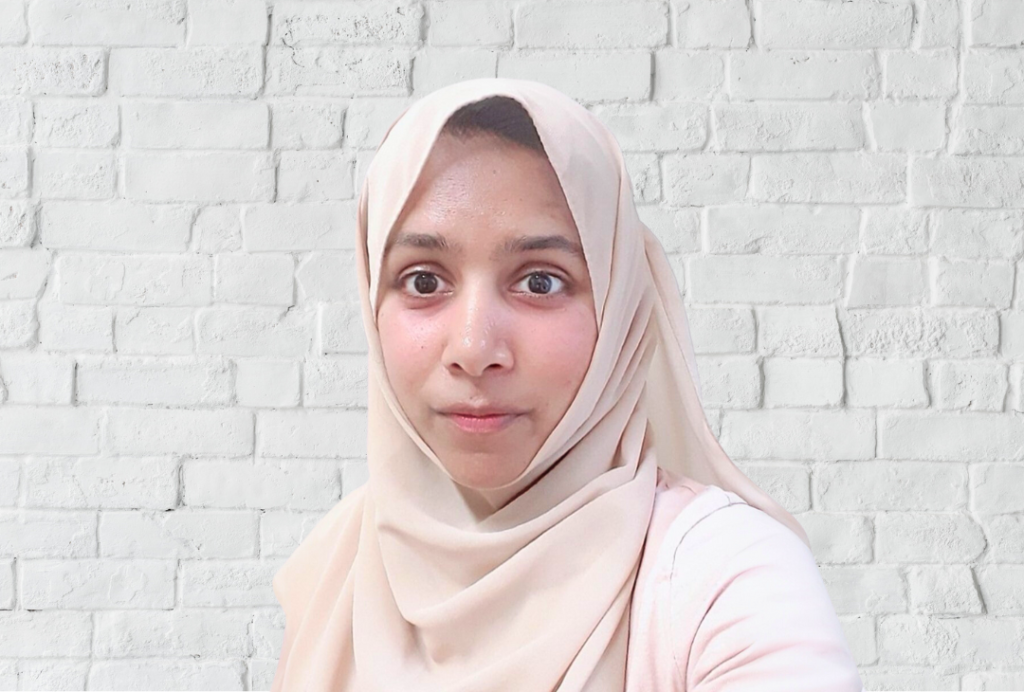 Woman looks at the camera. She is wearing a light pink hijab.