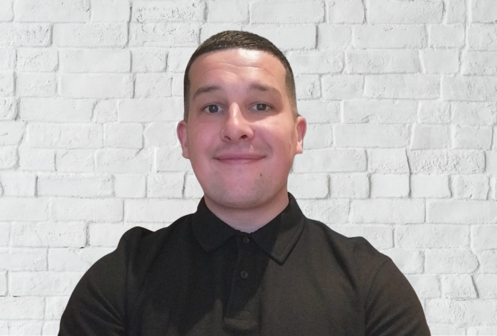 Man smiles at camera wearing a black tshirt against a block background.