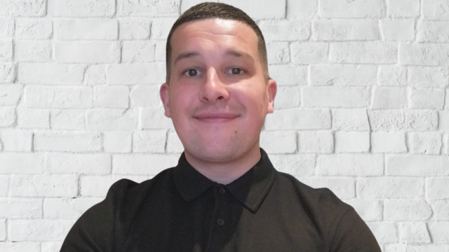 Man smiles at camera wearing a black tshirt against a block background.