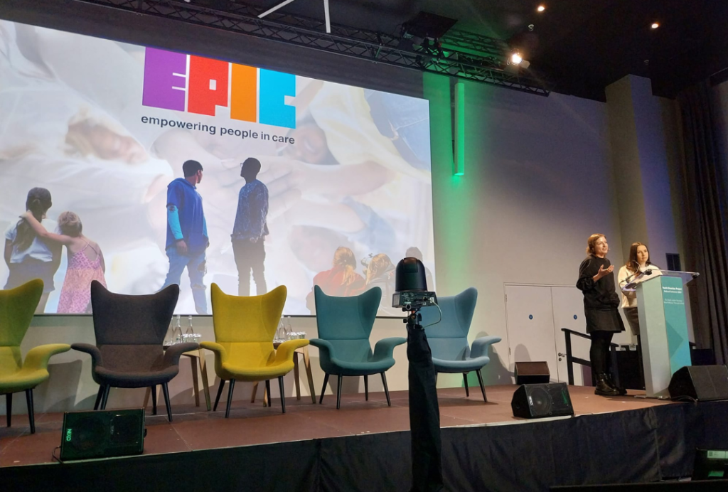 A member of our Youth Council and member of EPIC staff giving a presentation at a podium. An EPIC Presentation is on a large screen and there is five chairs in front of it.