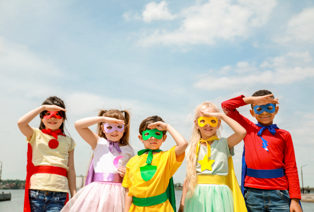 A group of children dressed as superheroes, with masks and capes, raise their hands to their heads.