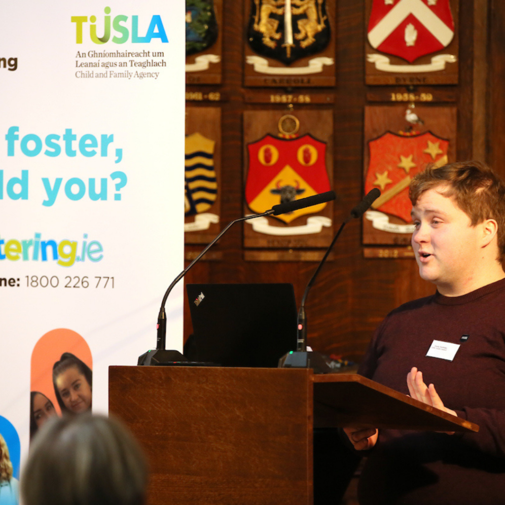 Young person speaking at a podium with a banner that has the Tusla logo.