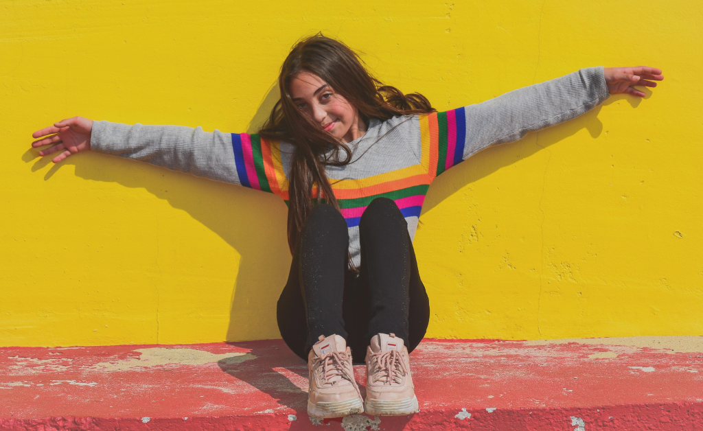 Teenage girl wearing a stripy rainbow jumper with her arms outstretched against a yellow wall.