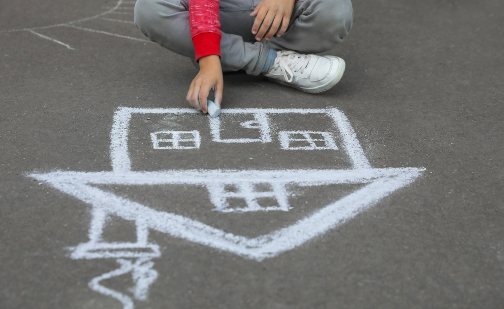 A child sits on the pavement and draws a house in white chalk.