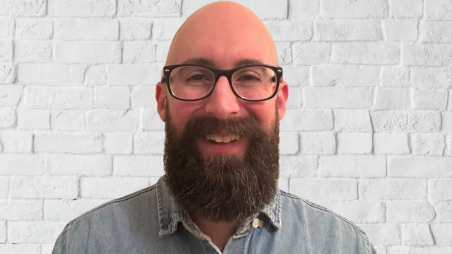 A man smiles at the camera. He is against a white wall background.