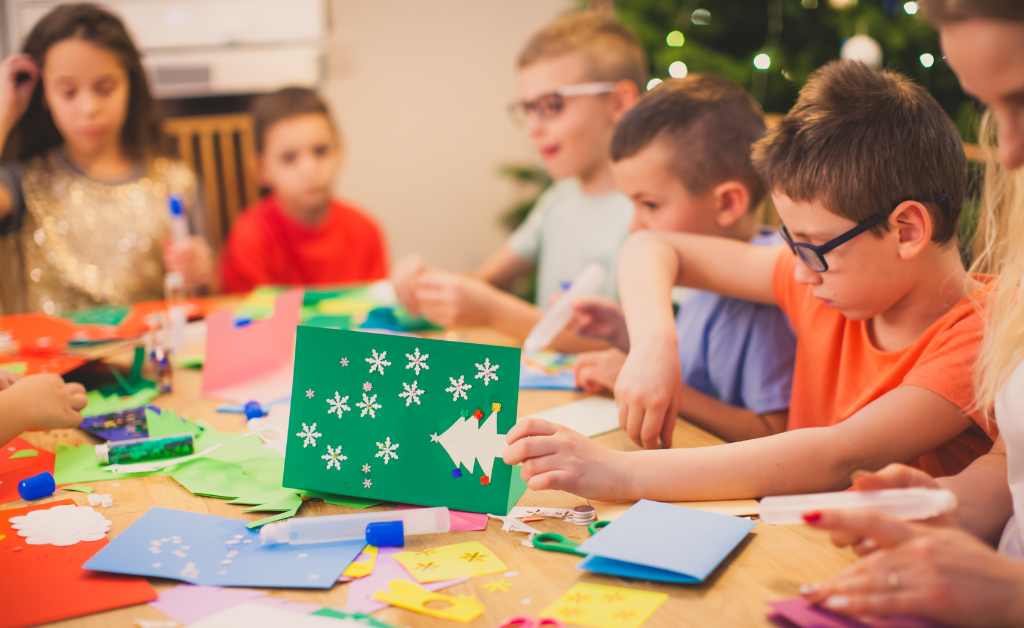 A festive photograph of young children making Christmas cards at a big table. There is lots of colourful card and craft materials on the table.