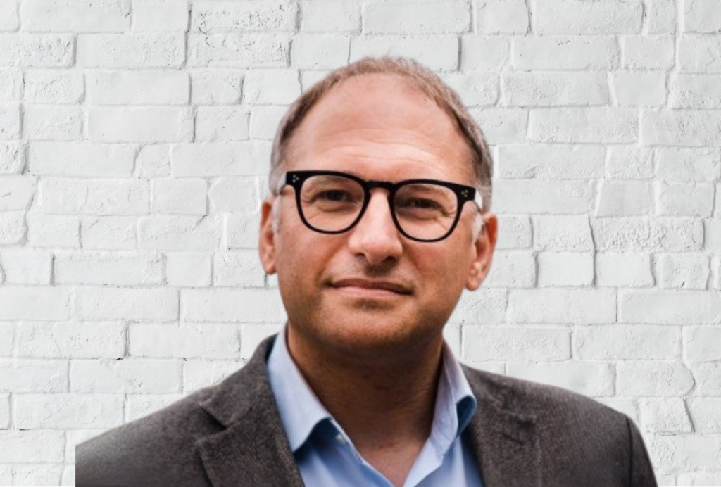 A man smiles at the camera. He is wearing glasses. The background is a white brick wall.