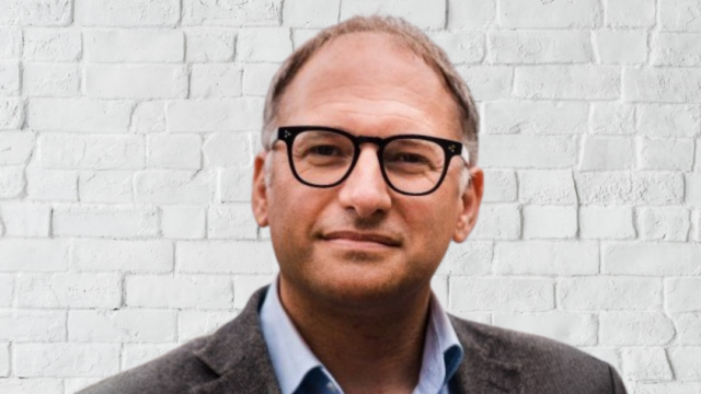 A man smiles at the camera. He is wearing glasses. The background is a white brick wall.