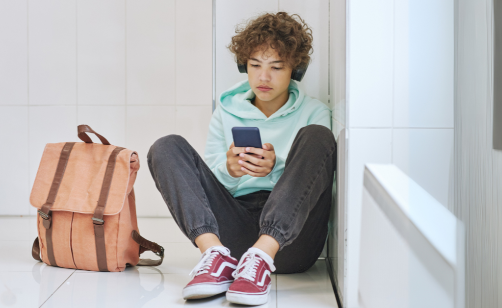 A young teenage boy sitting on the floor with his phone and a bag beside him